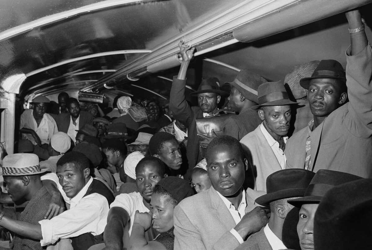 SOUTH AFRICA. 1960s. Africans throng Johannesburg station platform during late afternoon rush hour. Train accelerates with its load of clinging passengers. They ride like this through rain and cold, some for the entire journey. Inside, hands cling to a suitcase.